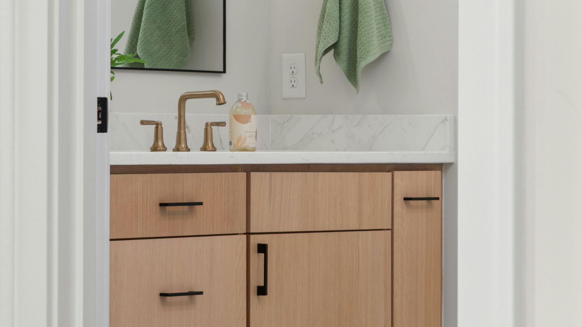 Bathroom vanity with light wood cabinets, white countertop, gold faucet, and green towels near a mirror
