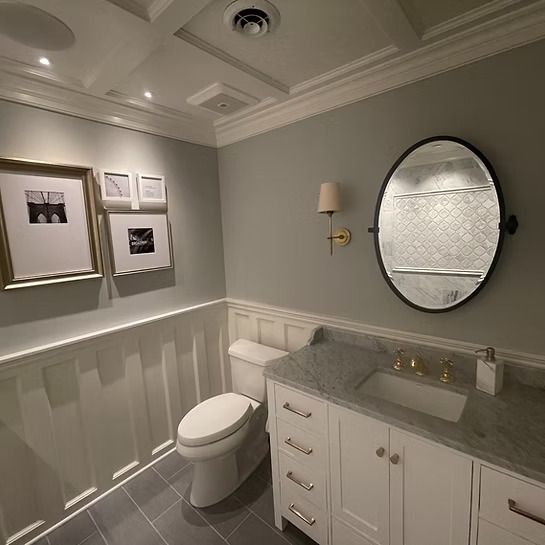 Elegant gray-and-white bathroom with vanity, round mirror, toilet, and framed wall art.