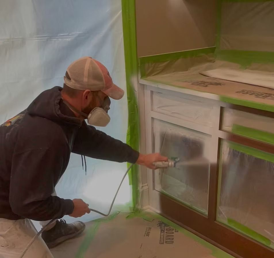 Person spray-painting cabinet doors while the surrounding area is masked with tape and plastic.