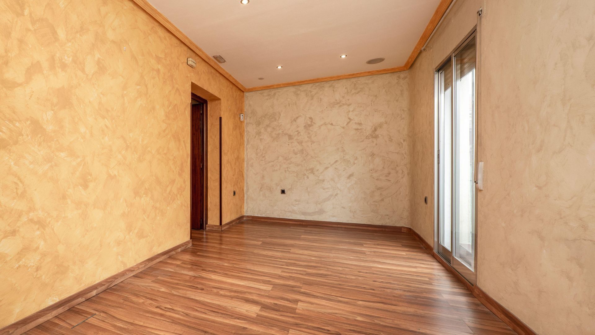 Empty beige room with wood flooring, recessed ceiling lights, and a sliding glass door on the right