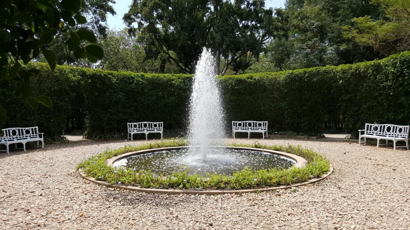 Fountain in a circular pond, surrounded by white benches and a hedge in a park.