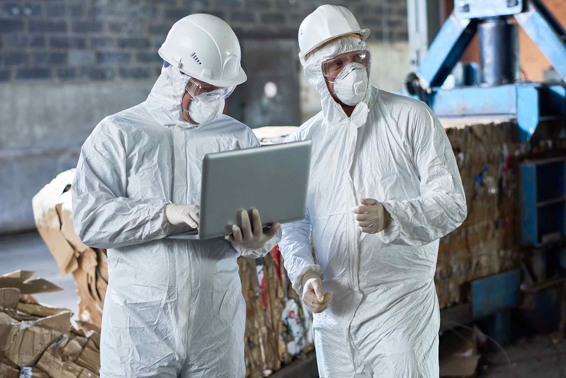Two men in protective suits are looking at a laptop in a factory.