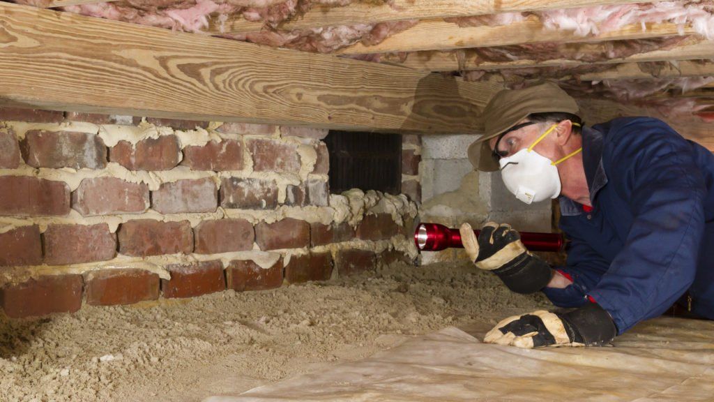 A man wearing a mask and gloves is looking under a brick wall with a flashlight.