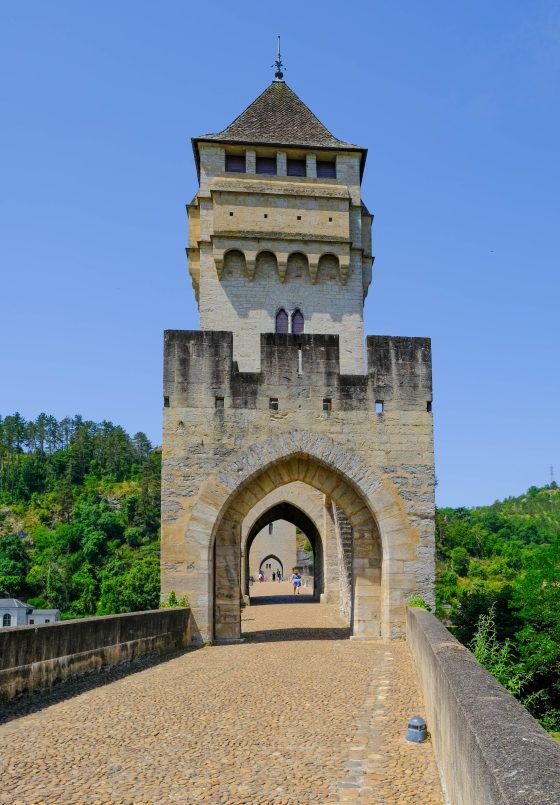 Stone gate tower with arched passage and tiled roof on a cobblestone bridge under a blue sky