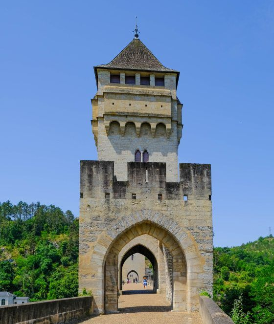 Stone gate tower with arched passageway on a bridge, against a blue sky and green trees