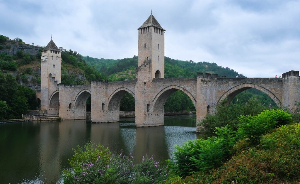 Stone bridge with two towers spanning a calm river, set in a green forested landscape under cloudy skies