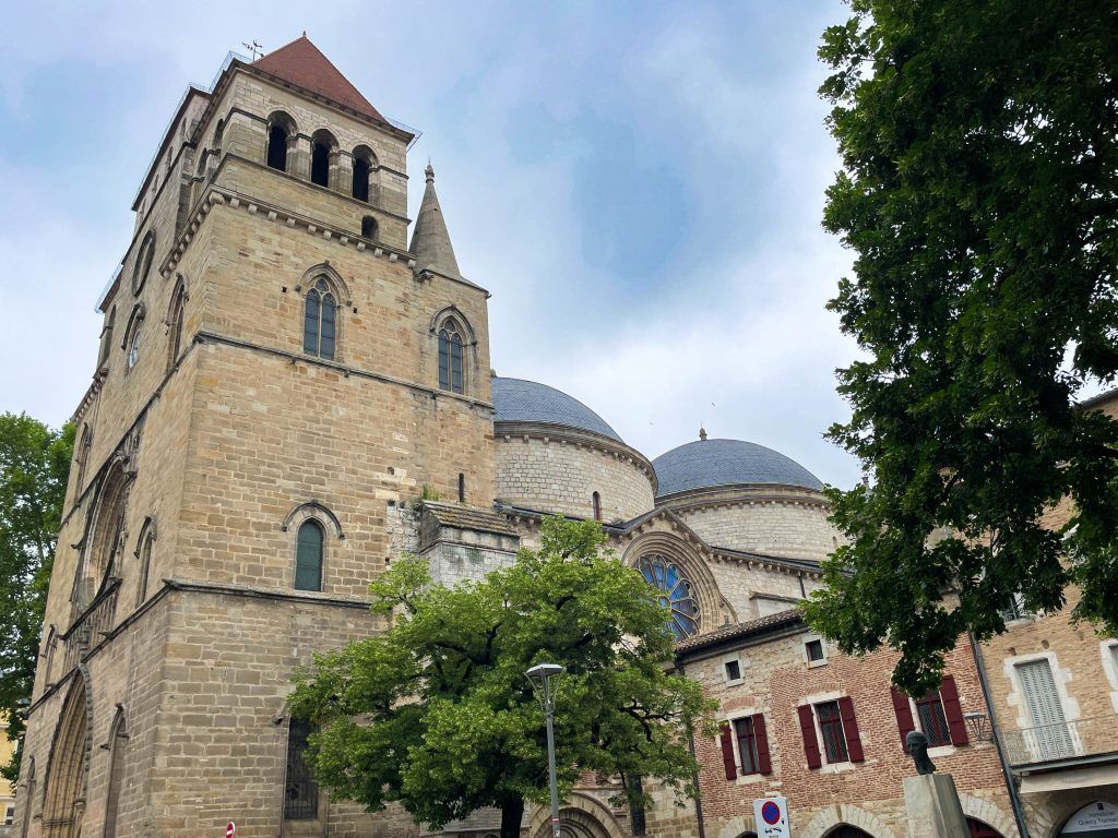 Stone church tower and domed roof beside a red building, framed by trees under a cloudy sky