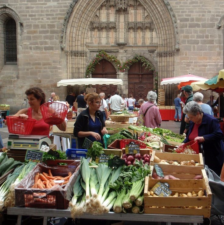 Outdoor market stall with fresh vegetables and shoppers in front of a stone building