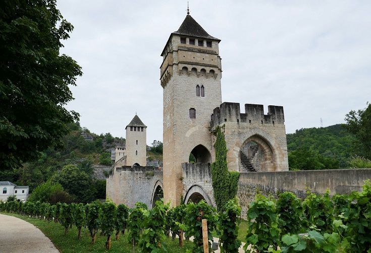 Stone castle tower and arched wall beside a vineyard, with trees and hills under a cloudy sky
