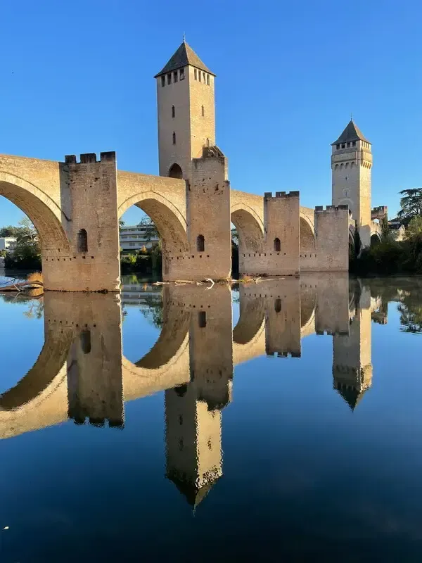 Stone bridge and towers reflected in calm water under a clear blue sky
