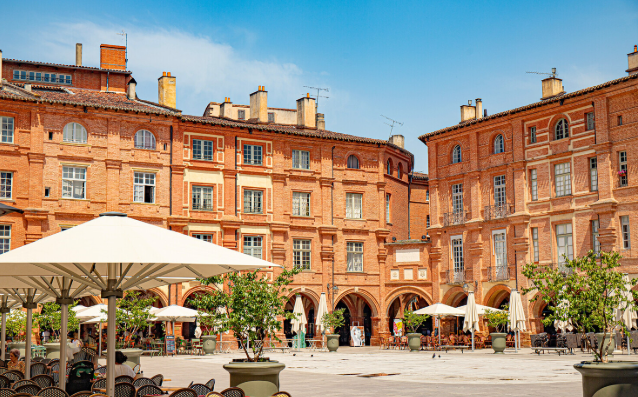 A square with umbrellas and chairs in front of a brick building in Toulouse, Fleur de Lys Retreats.