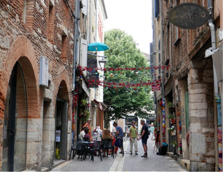 Narrow brick alley with hanging flowers, café tables, and people walking under archways