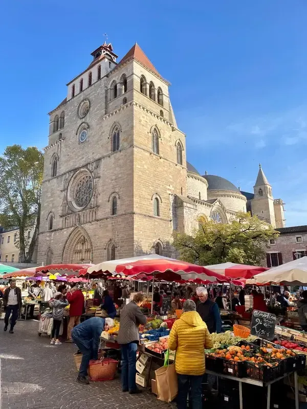 Outdoor market beside a stone church tower under a blue sky, with red umbrellas and shoppers browsing stalls