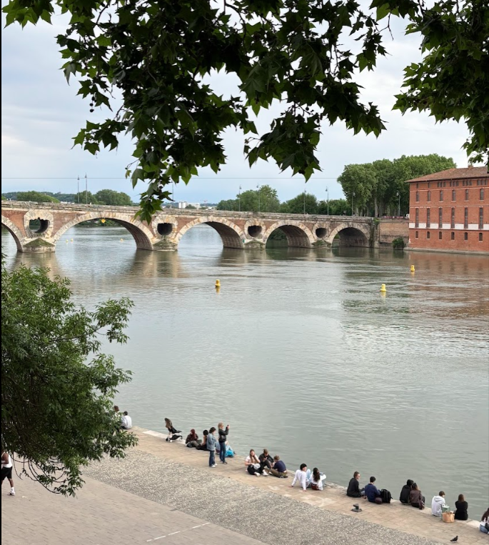 A group of people sit on the shore of a river with a bridge in the background