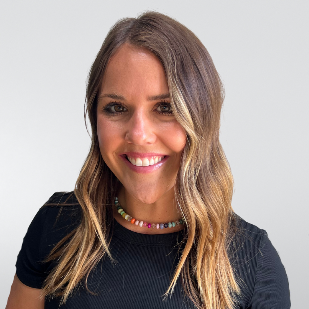 A smiling person with long wavy light brown hair wearing a black shirt and a colorful necklace against a gray backdrop.