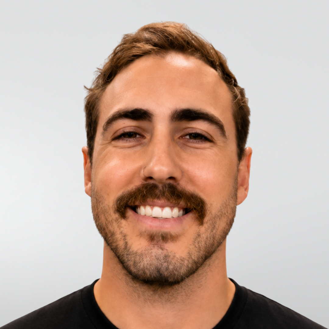 A person with brown hair and a beard, smiling at the camera, wearing a black shirt against a plain white background.