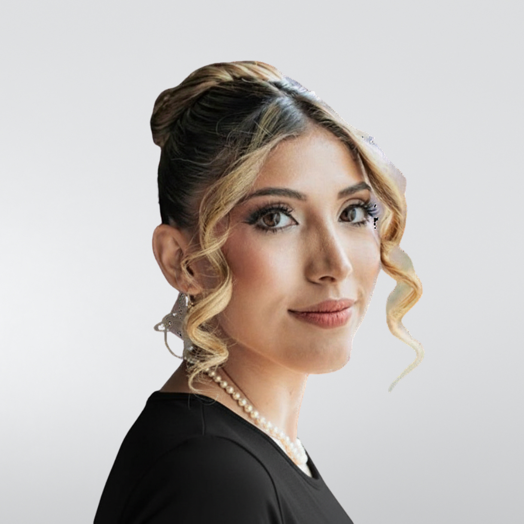 A person with hair styled in an updo with curled face-framing strands, wearing a pearl necklace against a plain background.