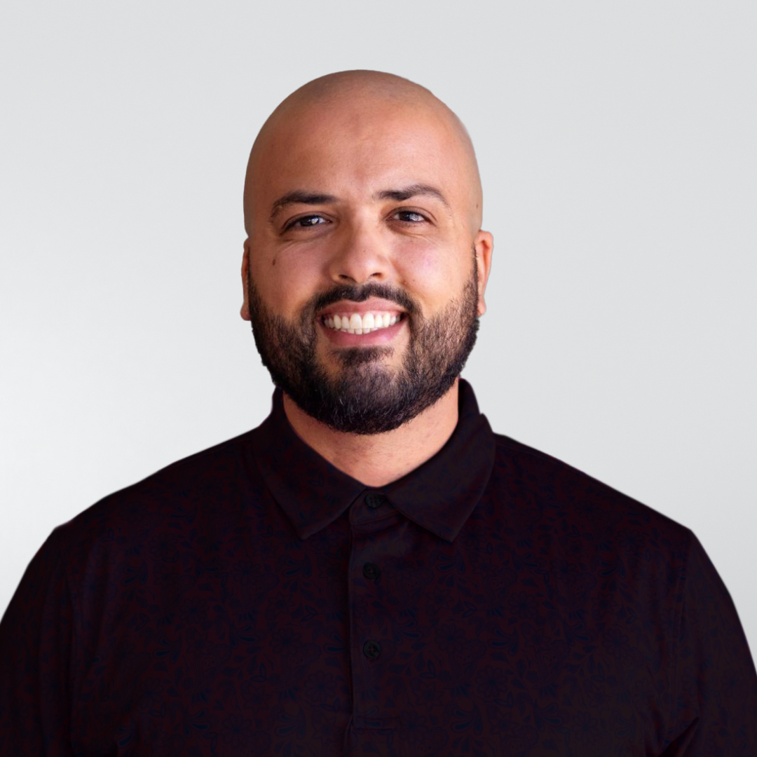 A smiling person with a shaved head and a dark beard, wearing a black collared shirt against a light grey background.