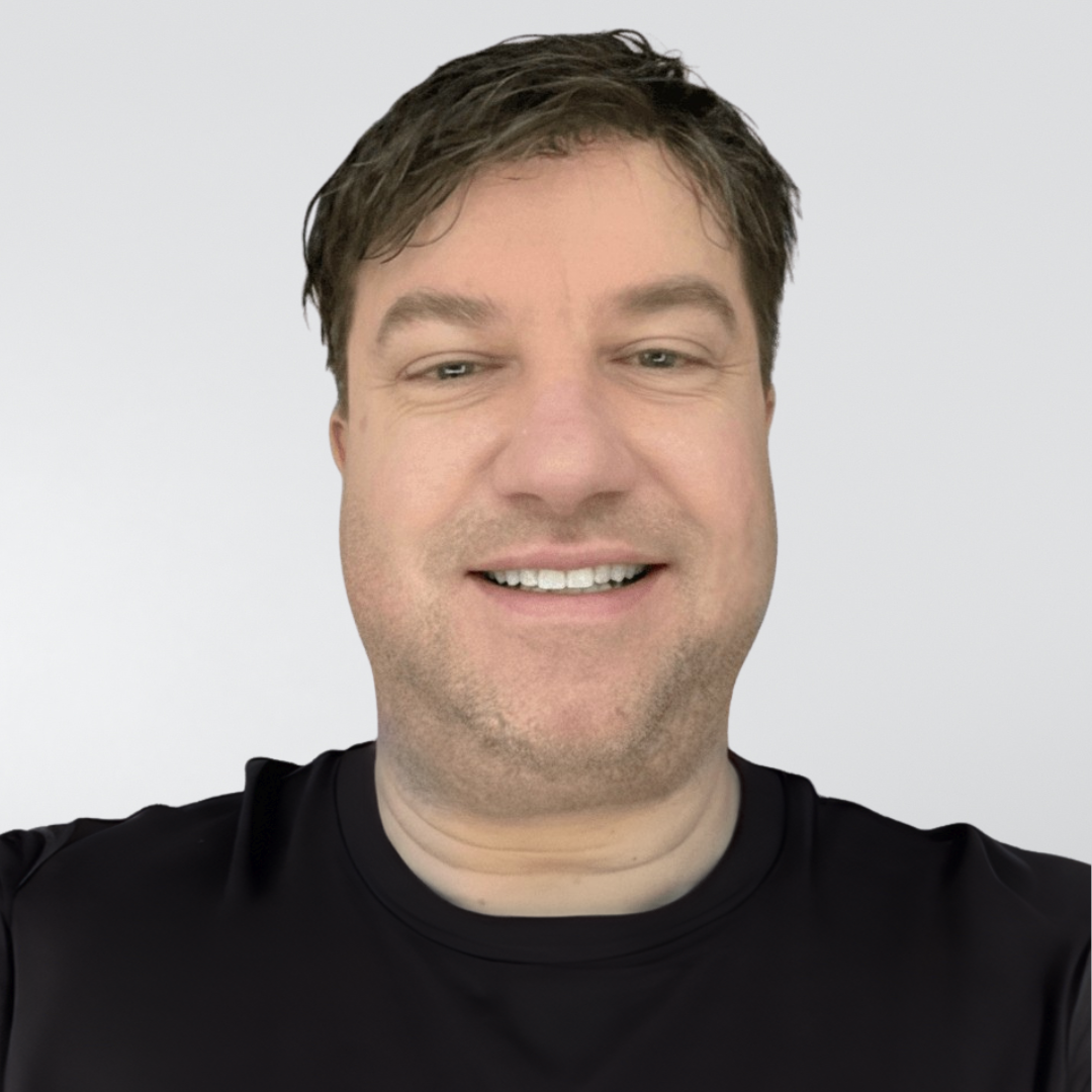 A person with short brown hair and a light stubble, smiling at the camera while wearing a black t-shirt against a grey wall.