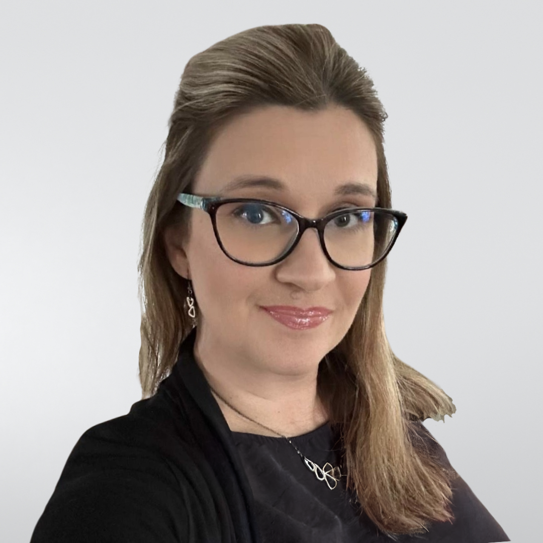 A person with long blonde hair and glasses wearing a black top smiles against a neutral gray background.