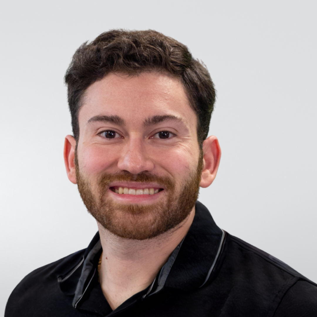 A man with dark hair and a beard, smiling at the camera, wearing a black polo shirt against a light gray background.