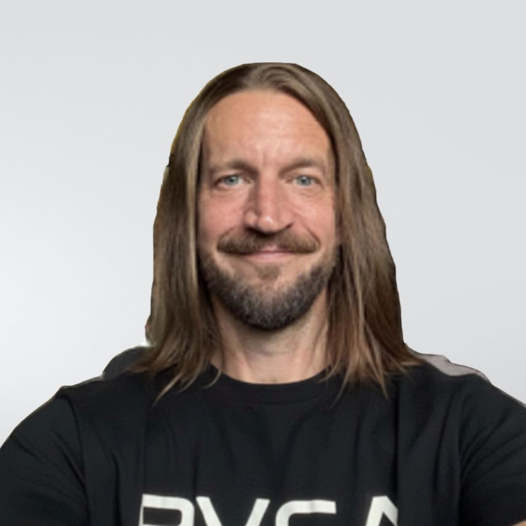 A person with long brown hair and a beard, wearing a black t-shirt with white text, smiling against a light gray background.