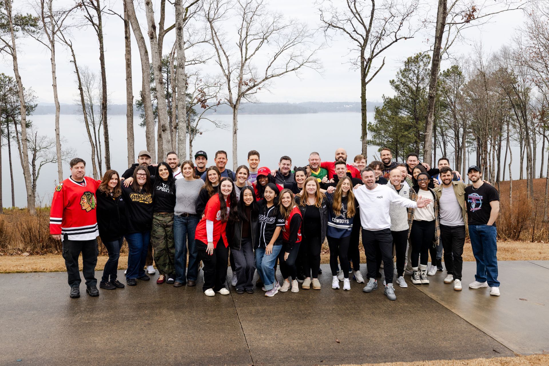 Large group posing outdoors on a wet road, with bare trees and a cloudy sky in the background.