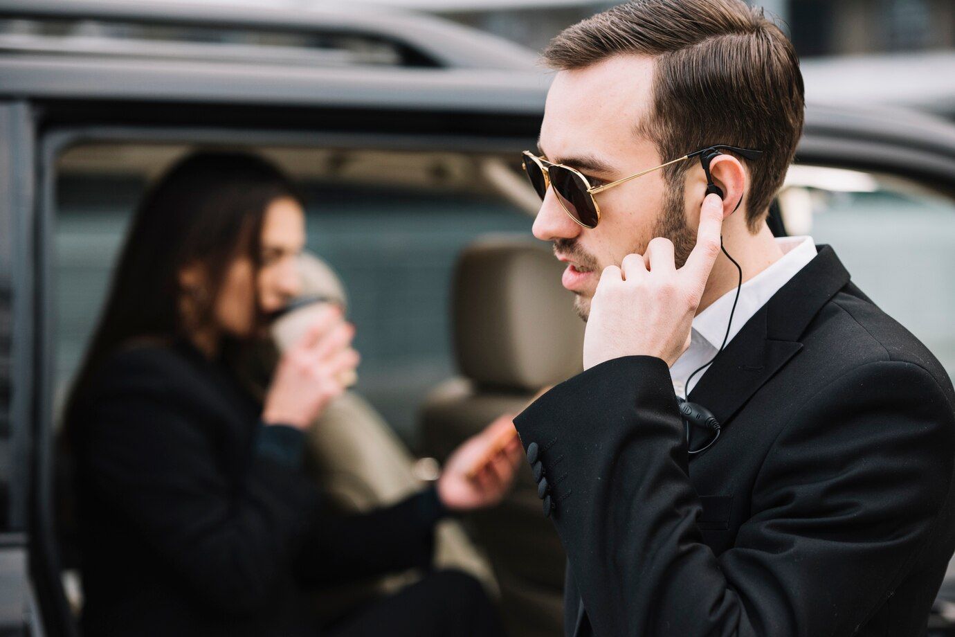 A man in a suit is talking on a cell phone while a woman drinks coffee in a car.