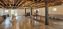 An empty event space with a concrete floor, exposed ceiling beams, string lights, and small cocktail tables.