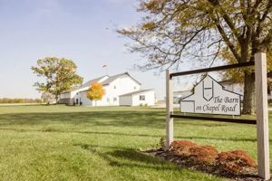 A white barn sits on a grassy field behind a sign that reads, 