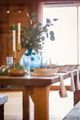 A wooden dining table set with white plates, a blue glass vase of greenery, and tall brass candlesticks.