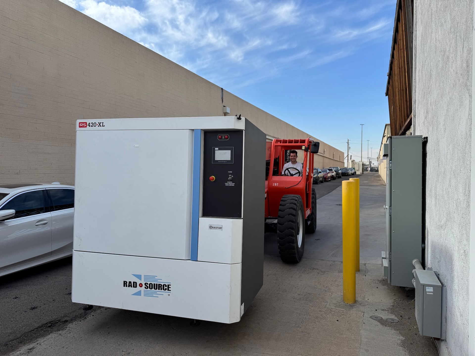 White utility enclosure beside a red truck in a narrow alley, with yellow bollards and a wall-mounted box.