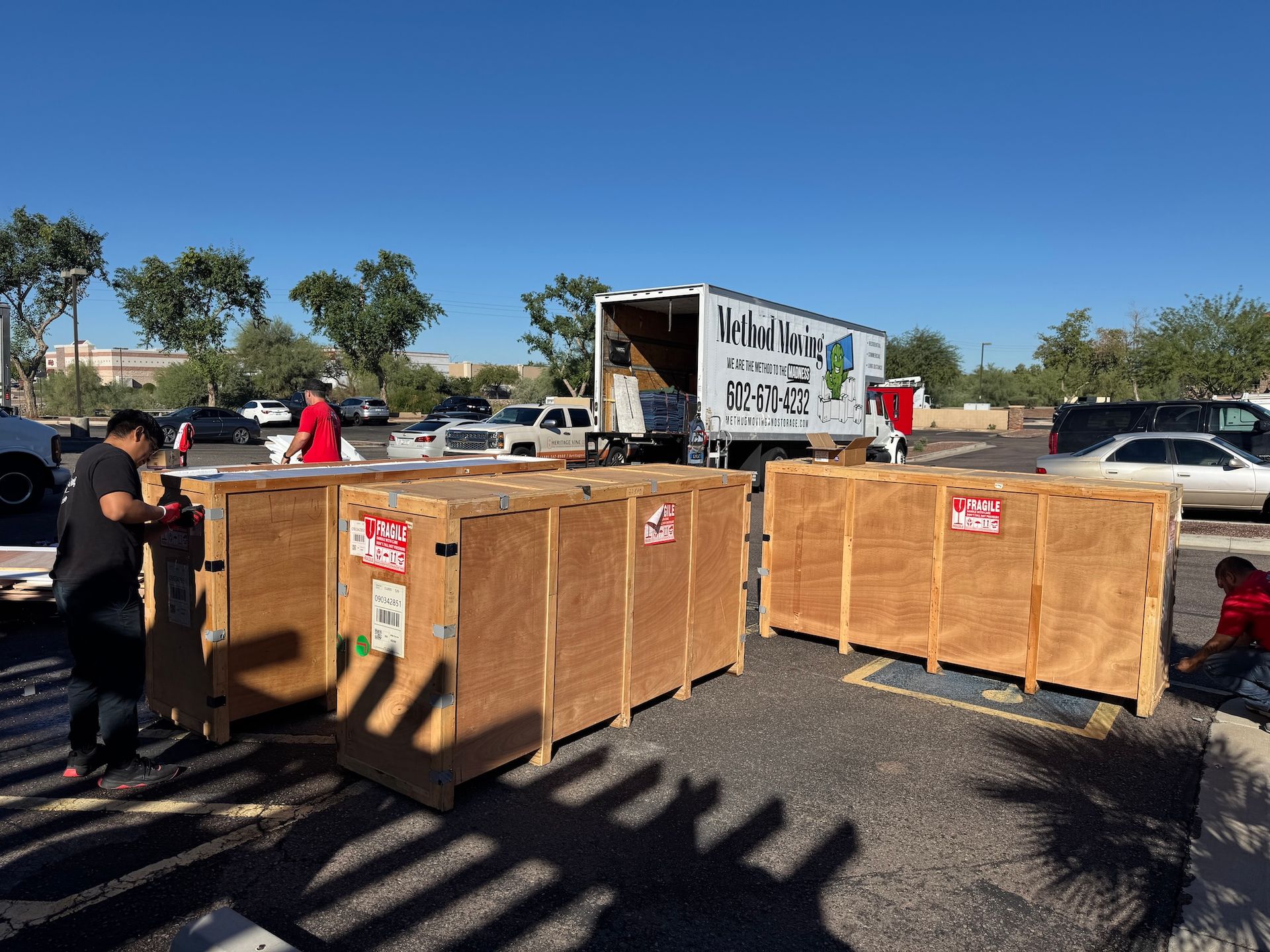 Large wooden crates being unloaded from a truck in a sunny parking lot, with a worker nearby.