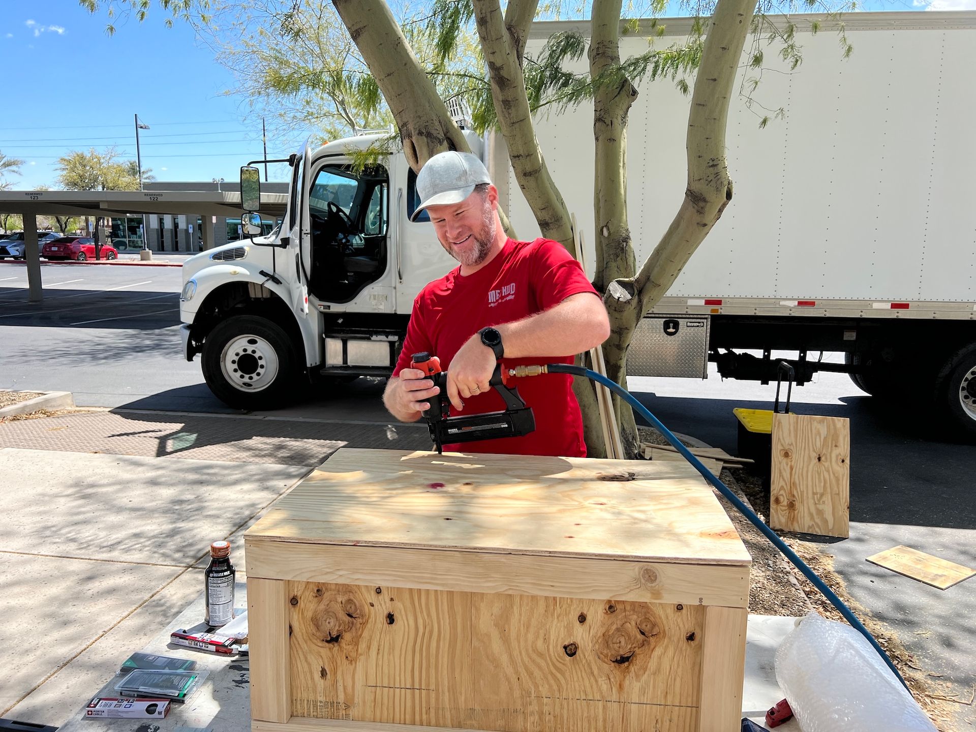Worker in red shirt using a drill on a wooden crate outdoors near a truck