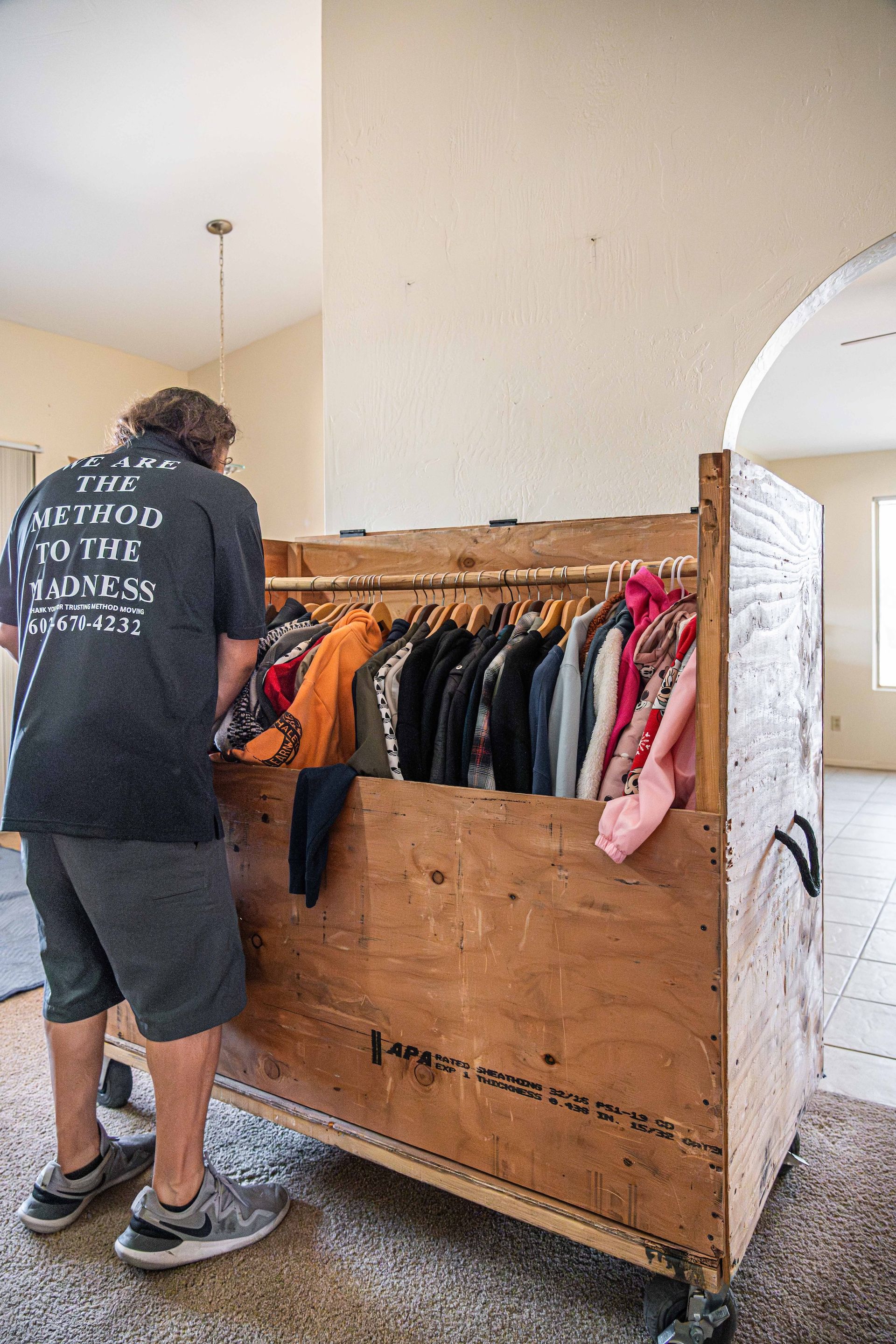 Person browsing clothes on a wooden rack in a home interior with carpet and arched doorway.