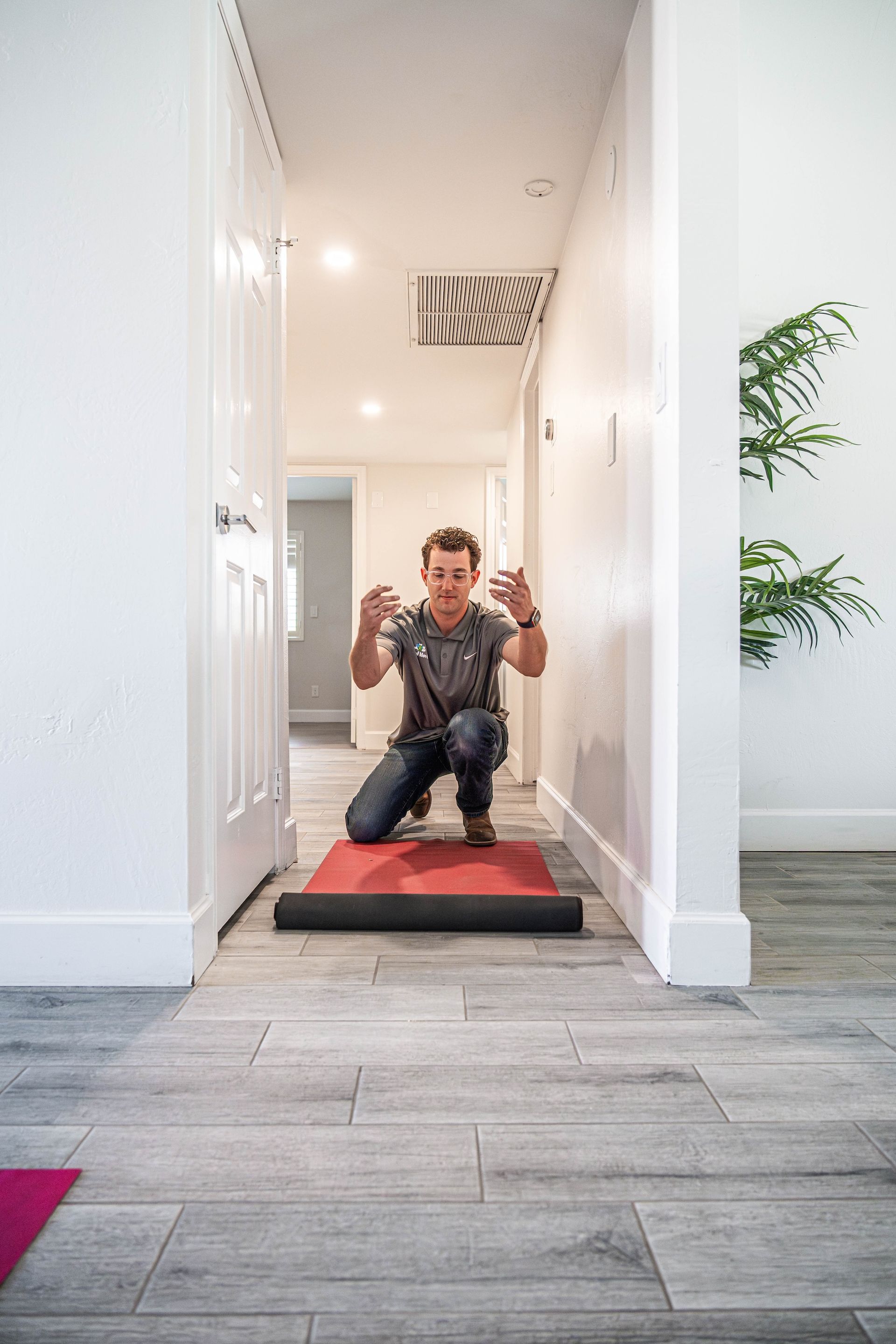 Person kneeling on a red mat in a bright hallway, posing with hands raised toward the camera.