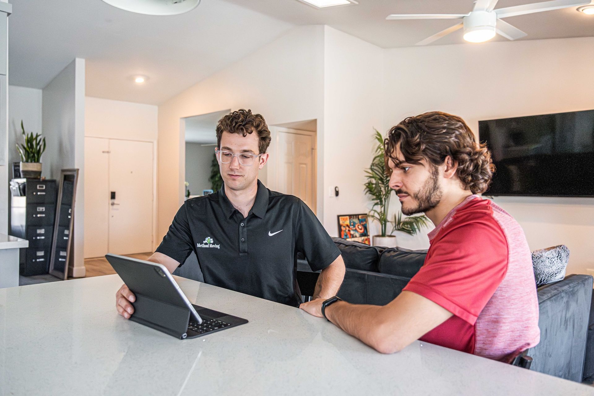 Two people sit at a kitchen counter looking at a laptop in a bright living room.