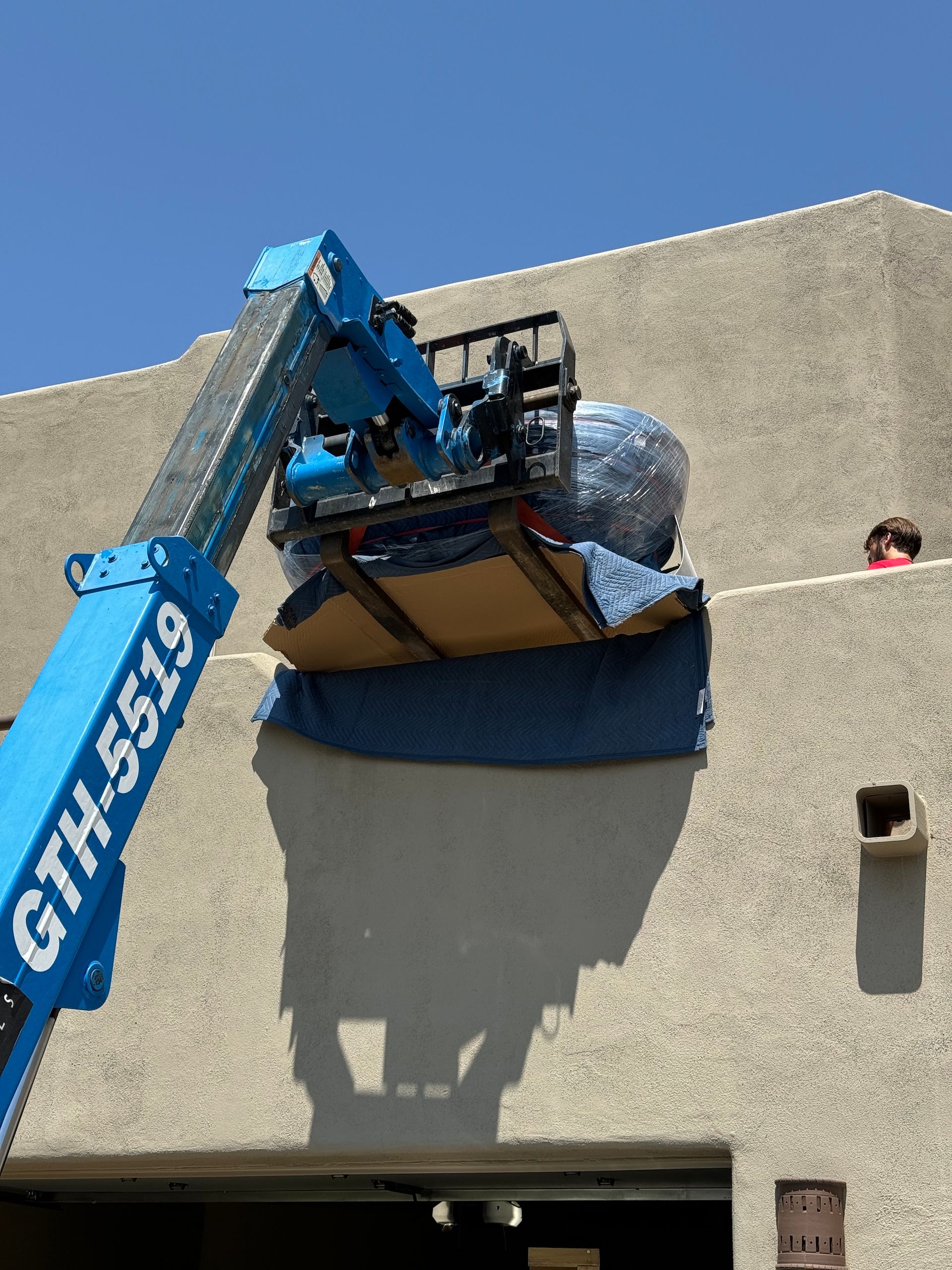 Blue GTH-5519 lift hoists a wrapped item over a beige wall under a clear sky