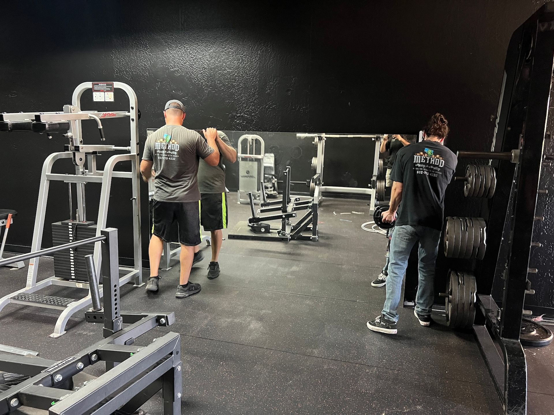 People in a gym near weight machines and a black wall, one standing by a mirror and another beside a rack