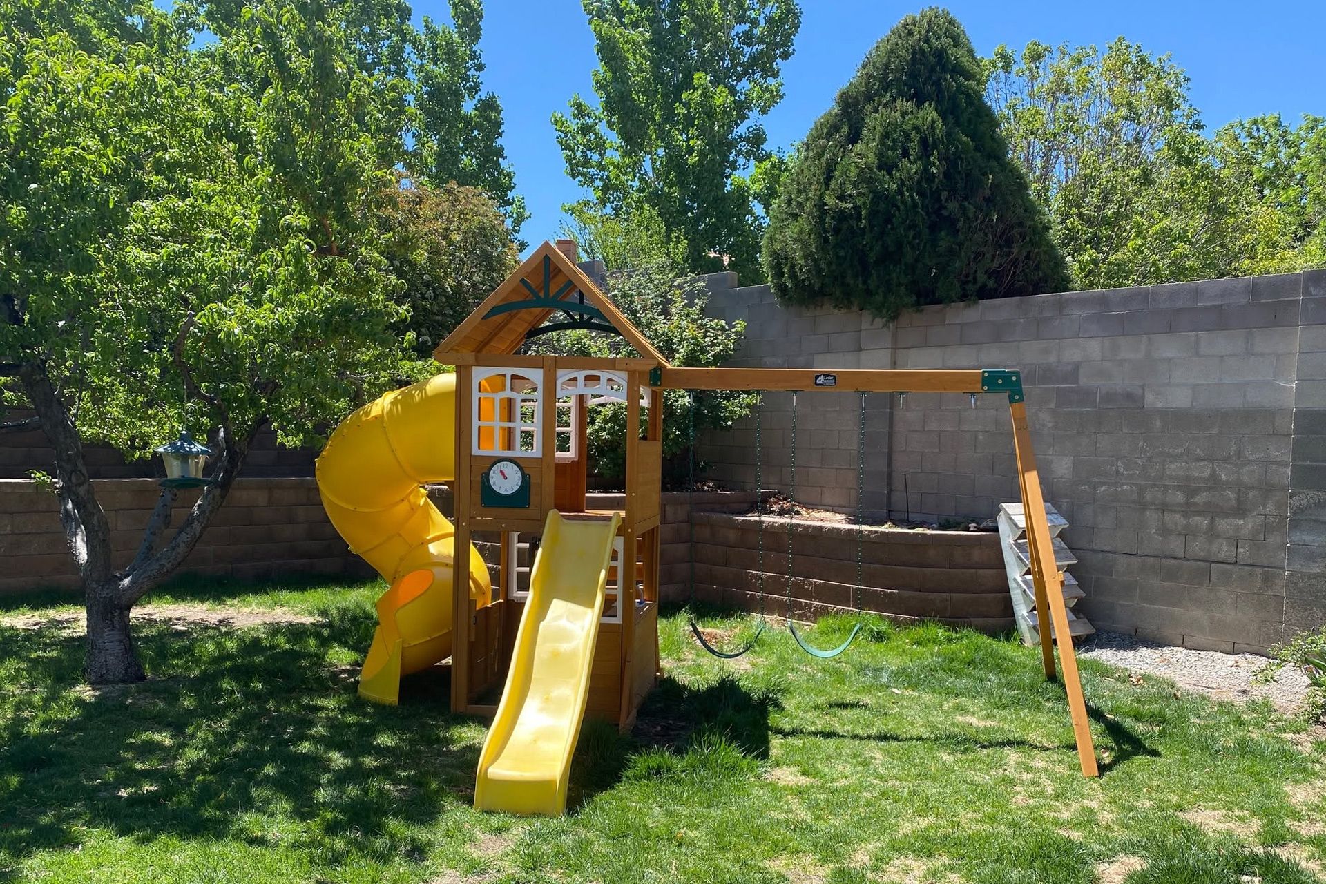 Yellow playground slide and wooden play structure in a backyard, with trees and a concrete wall.