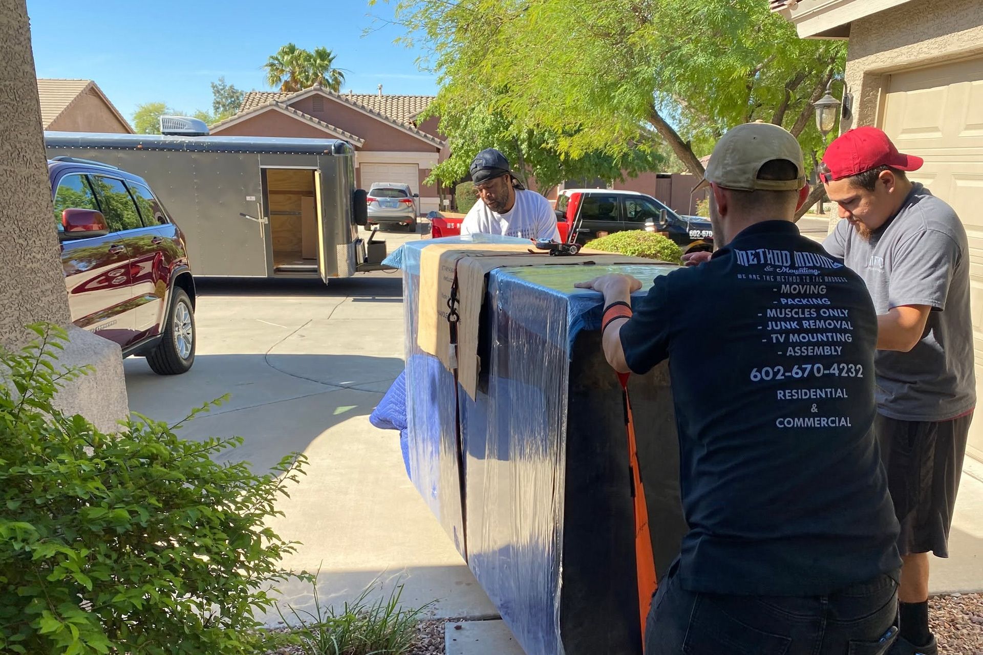 People moving a large appliance wrapped in blankets down a suburban driveway.