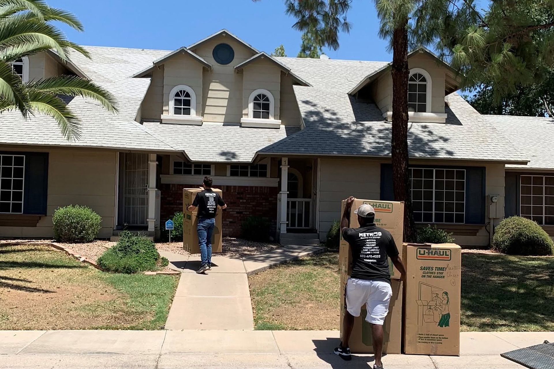 Two people carrying boxes stand outside a suburban house with a front walkway and palm tree.
