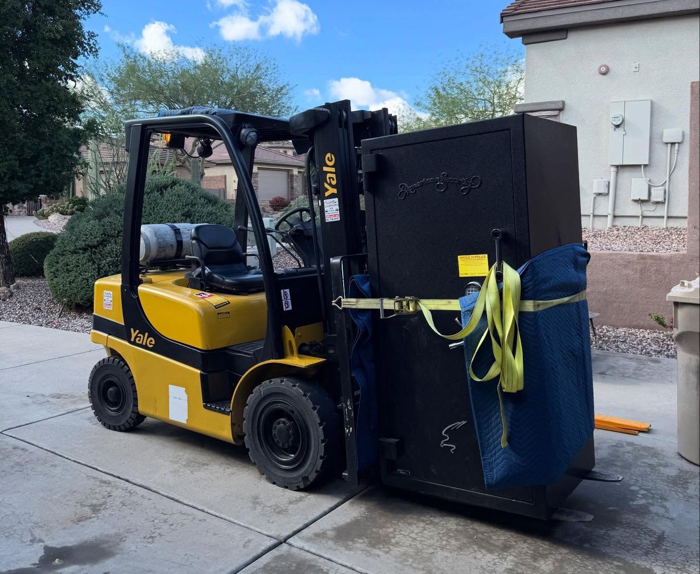 Yellow forklift carrying a large black bin with blue cover and yellow straps on a driveway