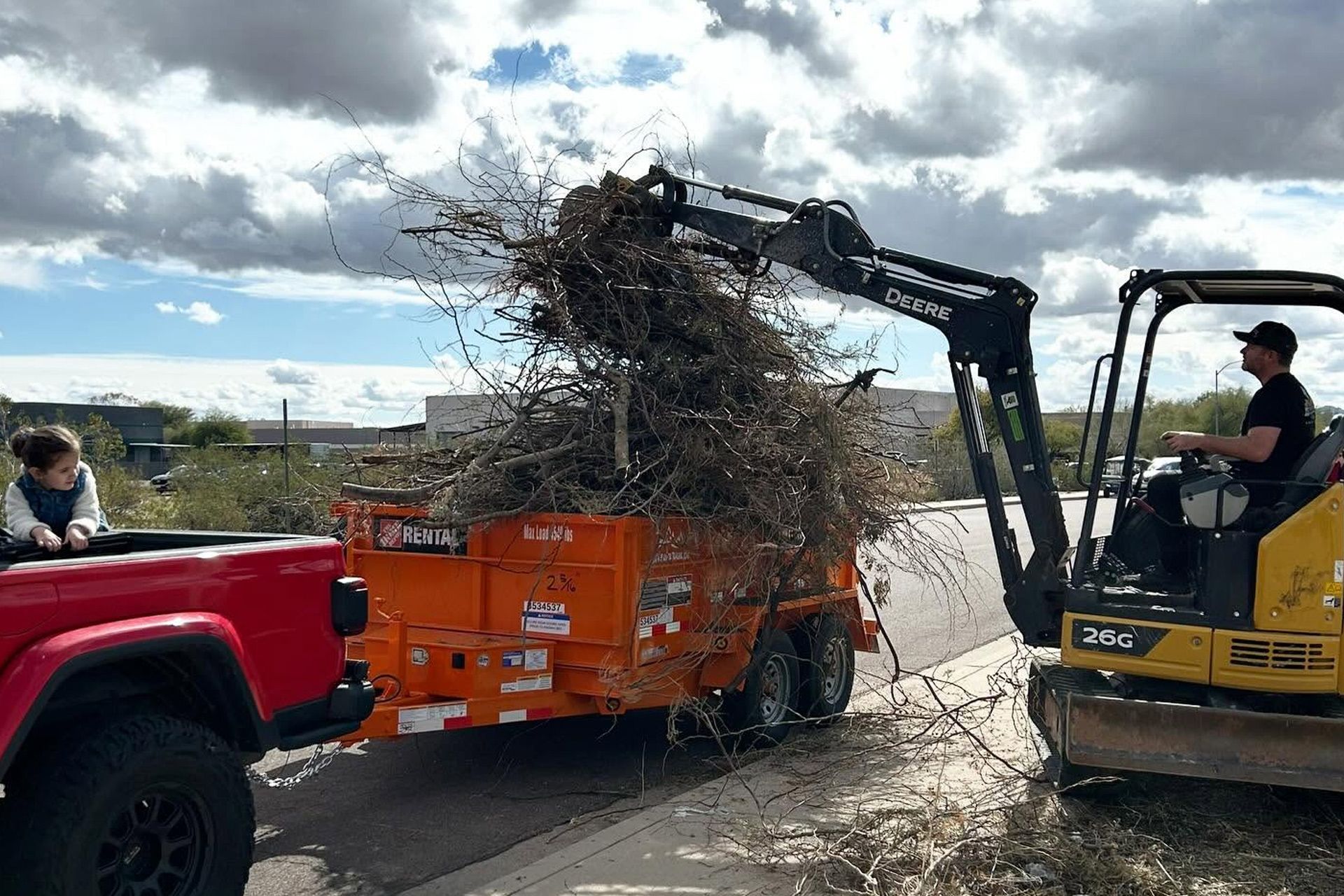 Excavator dumping brush into orange trailer while a red truck and worker stand nearby on a road