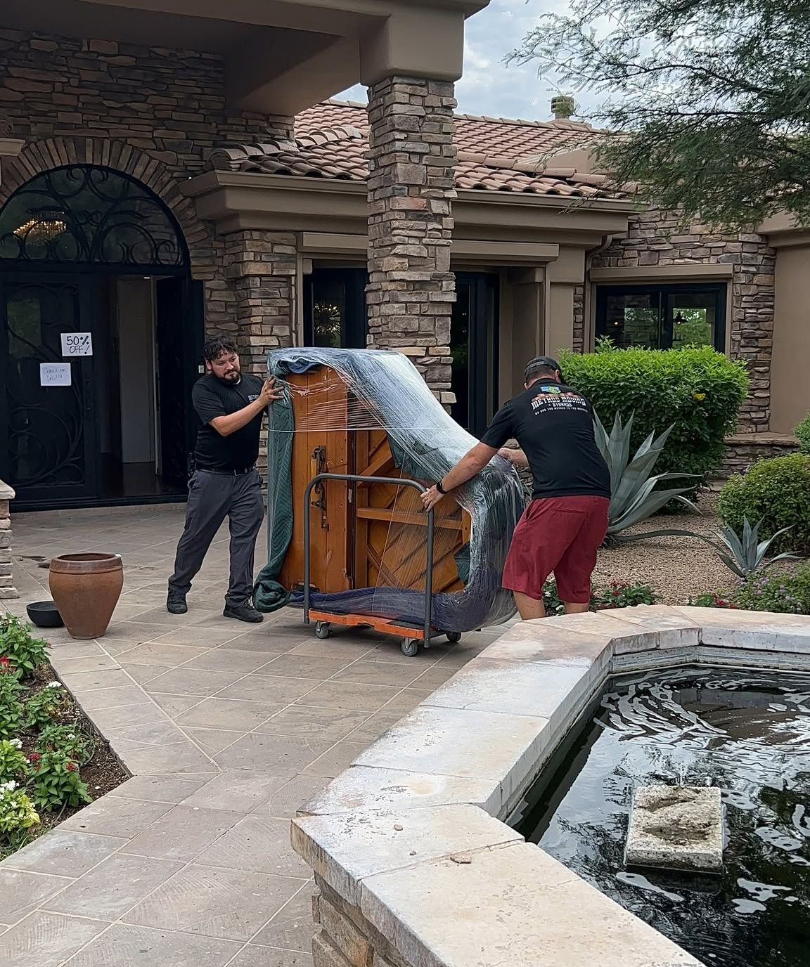 Two movers wrap a piano in plastic outside a house by a fountain