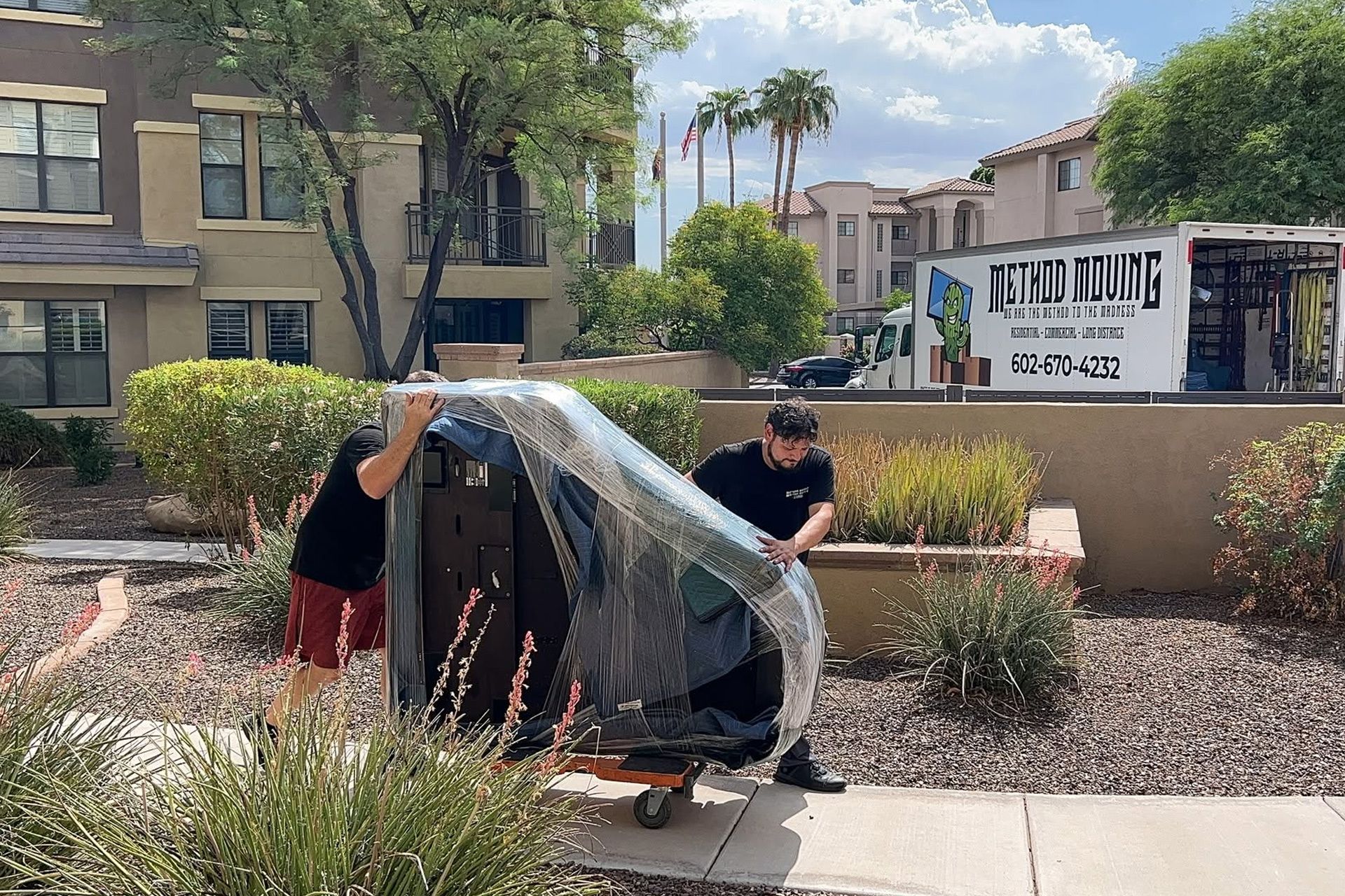 Two people load a black cart under plastic wrap on a sidewalk near apartment buildings
