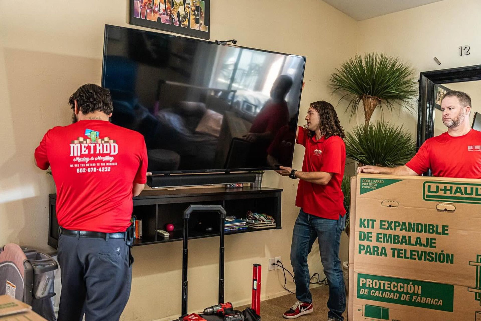 Three movers in red shirts lift a large TV in a living room with U-Haul boxes.