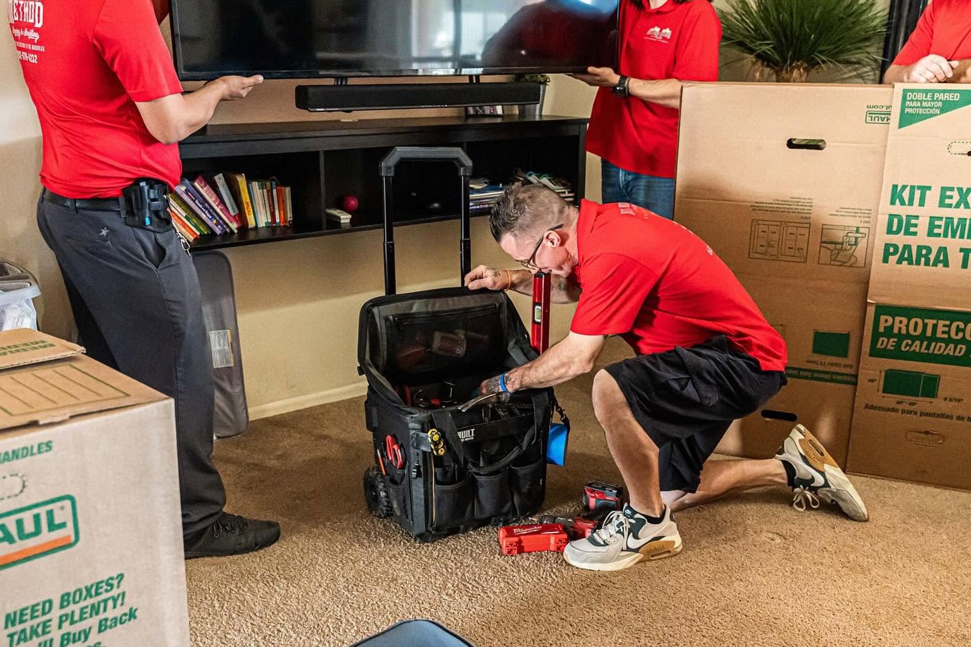 Workers in red shirts pack a large black tool bag in a store aisle beside boxes.