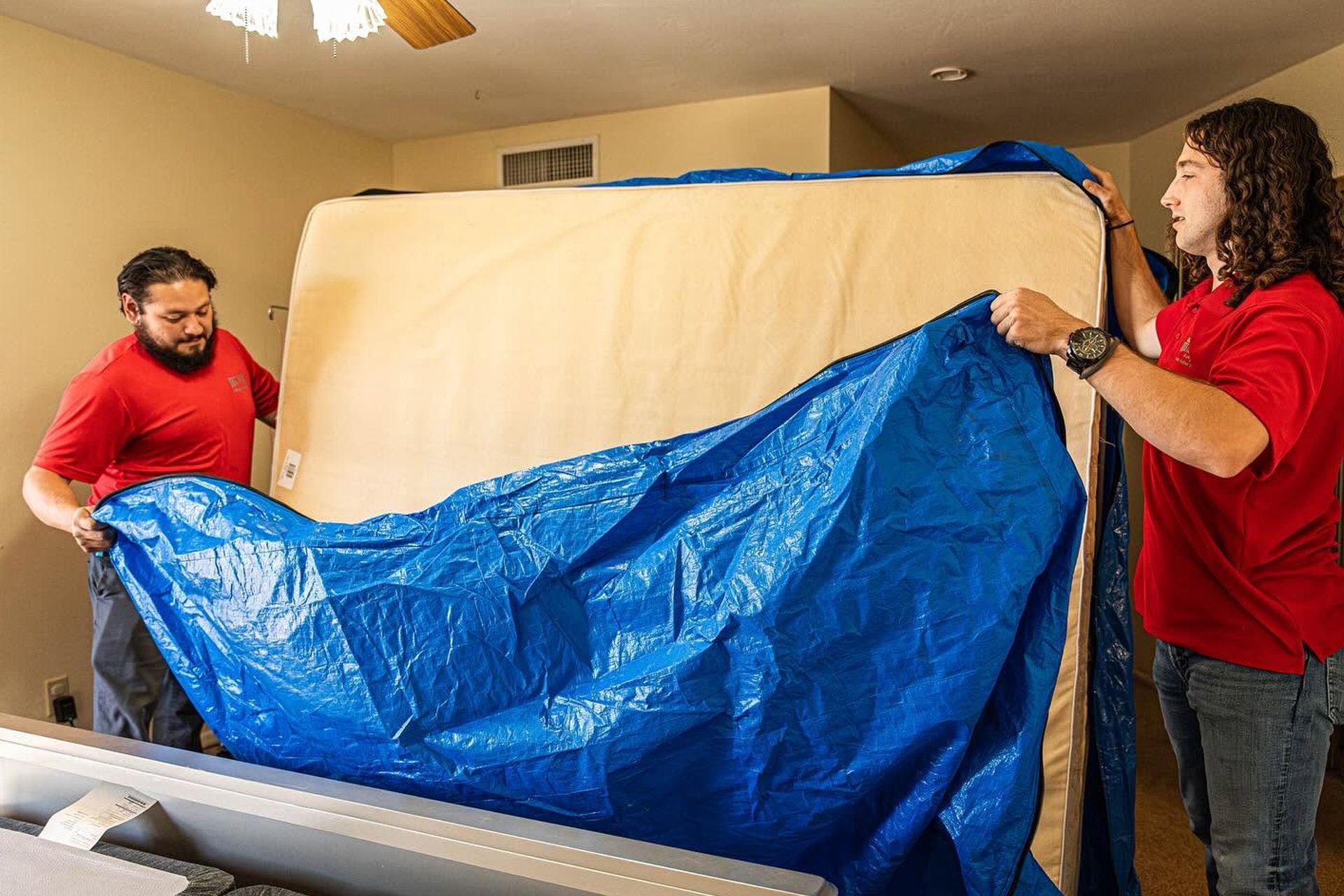 Two people in red shirts stretch a blue tarp over a large beige mattress indoors.