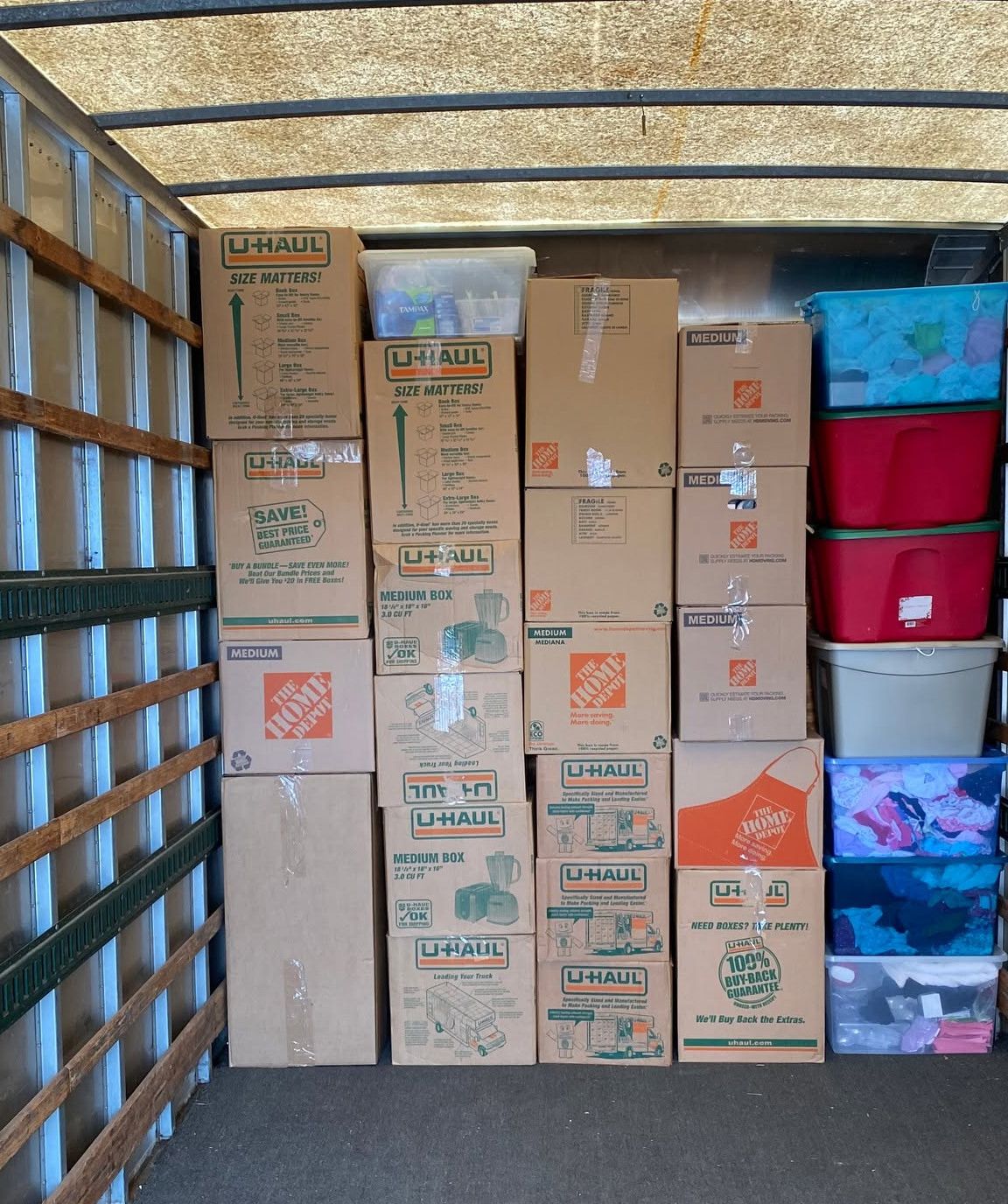 Cardboard boxes stacked in a moving truck beside plastic storage bins and a rolled rug
